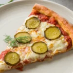 Close-up of a pickle pie pizza slice on a white plate, showcasing golden crust, garlic sauce, and vibrant dill pickles.